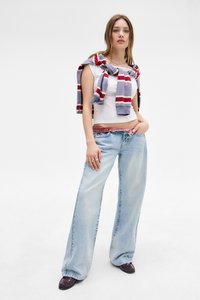 Jeans de pierna ancha azul claro con una textura desgastada, combinados con una blusa sin mangas blanca. Un suéter de rayas multicolores está atado alrededor de los hombros.