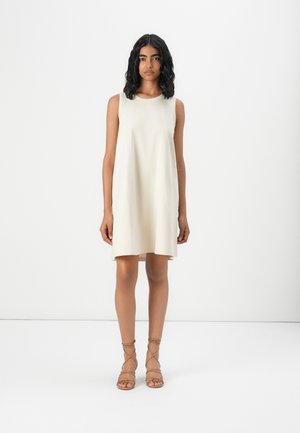 Femme aux cheveux bouclés foncés portant une robe crème sans manches et des sandales marron à brides, debout devant un fond blanc uni.
