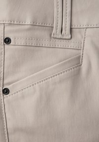 Beigefarbene Hosentasche mit verstärkten Nähten, zwei schwarzen Nieten und einer Gürtelschlaufe oberhalb des Taschenrands.