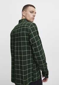 Cămașă verde cu carouri, realizată dintr-un material cu o textură moale, având linii albe formând un grid, cu închidere prin nasturi și mâneci lungi.