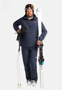 Skiausrüstung mit einer marineblauen wasserdichten Jacke und Hose, weißen Skiern mit schwarzen Bindungen sowie Winterhandschuhen, getragen mit Skischuhen.
