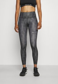 Sprawna osoba ubrana w czarne i szare legginsy Nike z nadrukiem logo oraz czarny stanik sportowy, stojąca na jednolitym tle.