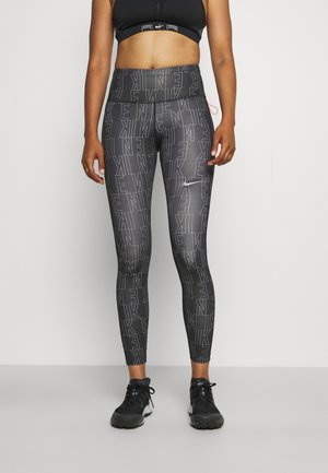Une personne en forme portant des leggings Nike noirs et gris avec un imprimé de logo sur l'ensemble et un soutien-gorge de sport noir, se tenant devant un fond uni.