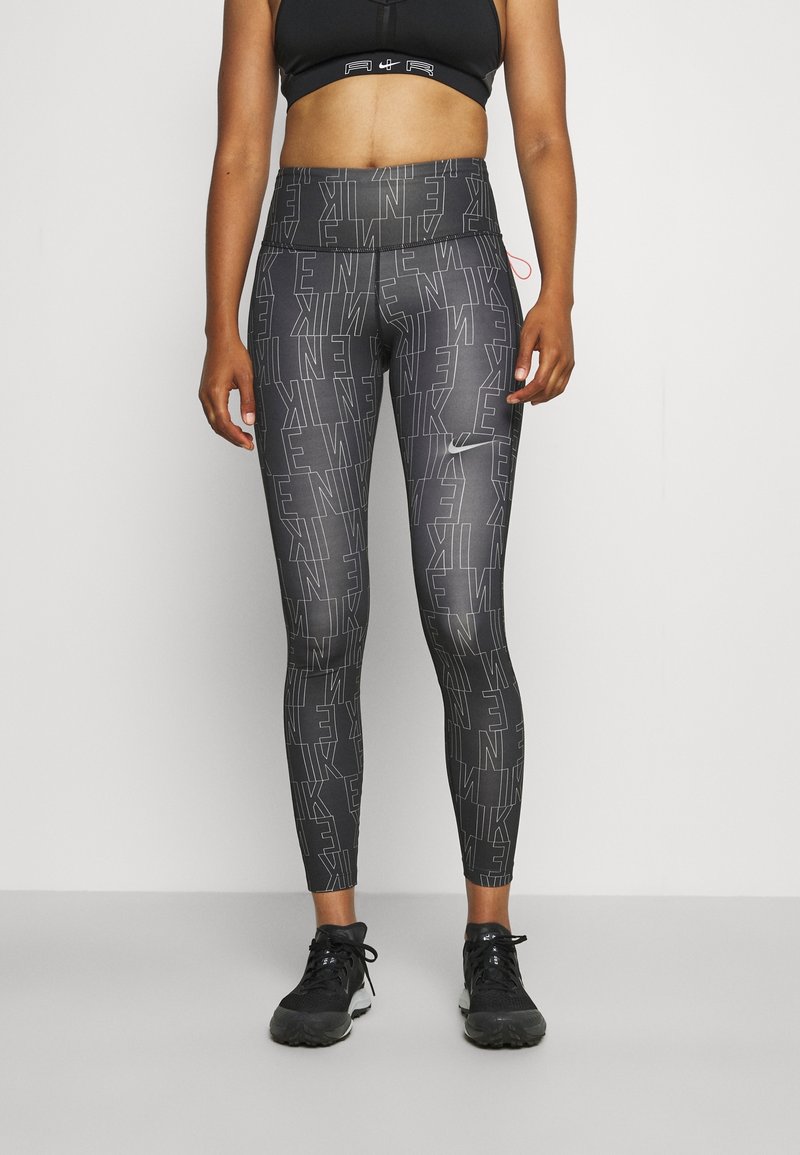Sprawna osoba ubrana w czarne i szare legginsy Nike z nadrukiem logo oraz czarny stanik sportowy, stojąca na jednolitym tle.