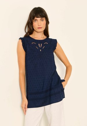 Woman wearing a navy blue sleeveless blouse with eyelet embroidery and ruffled shoulders, paired with white pants, standing against a plain background.