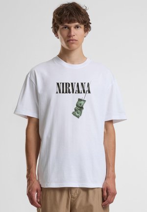 Jeune homme aux cheveux courts et bouclés porte un t-shirt blanc Nirvana avec un motif billet de dollar, debout devant un fond clair uni.