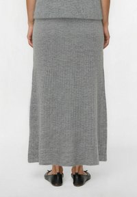 Vrouw in een geribbelde, enkellange grijze gebreide rok en zwarte leren ballet flats, gezien van achteren tegen een witte achtergrond.