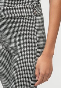 Leggings i sort-hvid houndstooth mønster med en tætsiddende pasform, der har en lille metallisk detalje nær taljen og en glat tekstur.