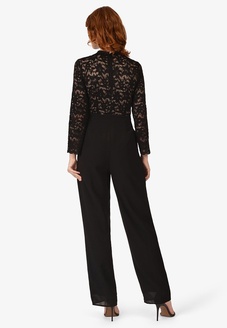 Kobieta z falującymi, czerwonymi włosami, ubrana w czarną, koronkową bluzkę z długim rękawem oraz czarne spodnie z wysokim stanem, odwrócona tyłem, stojąca na białym tle.