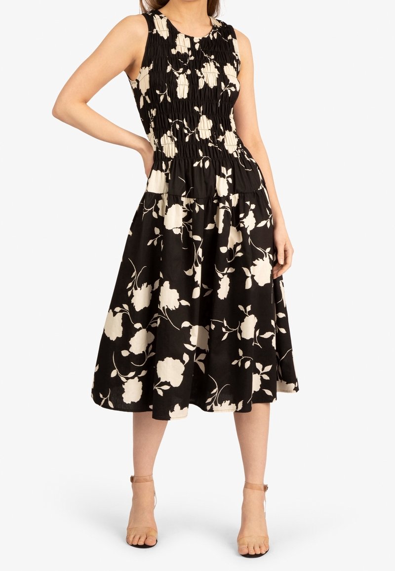 Femme portant une robe midi noire sans manches à imprimé floral blanc et corsage froncé, associée à des talons nude à bride à la cheville.