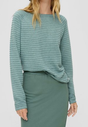Femme portant un haut vert côtelé à manches longues, rentré dans une jupe verte ajustée sur un fond uni.