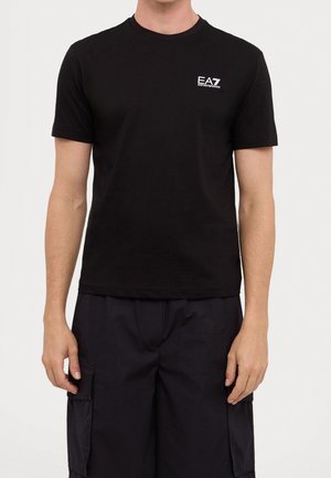 Mężczyzna w czarnej koszulce EA7 Emporio Armani i czarnych szortach cargo stojący na białym tle.