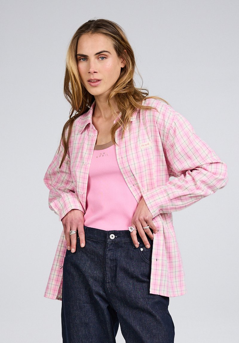 Sieviete ar gariem blondiem matiem, valkā rozā tanktopu, rozā rūtainu pogājamu kreklu, tumši zilas džinsas un gredzenus uz pirkstiem.