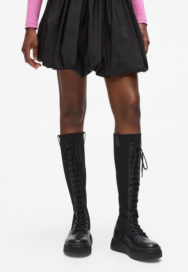 Personne portant des bottes montantes noires à lacets avec des semelles épaisses et une jupe noire bouffante, debout sur un fond blanc.