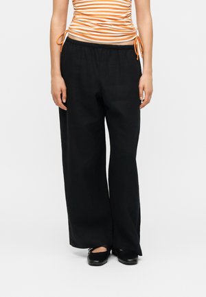 Vrouw met losse zwarte broek, oranje-wit gestreept gerimpeld topje en zwarte ballerina's, staand tegen een effen achtergrond.