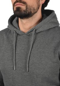 Sweat à capuche gris en mélange de coton, avec un cordon de serrage et des œillets en métal, offrant une texture douce. Gros plan sur l'encolure et la capuche.