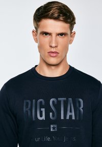 Jaunas vyras trumpais rudos spalvos plaukais, dėvintis tamsų džemperį su užrašu „BIG STAR“ ir žvaigždės logotipu priekyje.