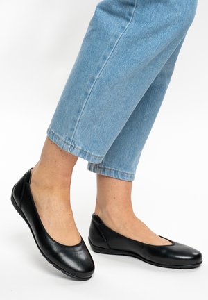 Füße mit schwarzen Ballerinas aus Leder, kombiniert mit hellblauen, kurz geschnittenen Jeans, vor weißem Hintergrund.