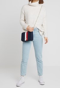 Bolso cruzado azul marino con un acento de franjas rojas y blancas, material suave, forma rectangular y una cadena como strap, combinado con un suéter color crema y jeans claros.