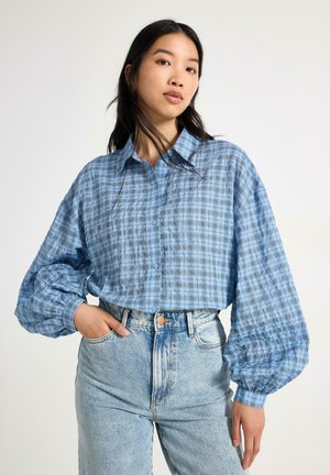 Kvinde iført en blå ternet bluse med puffede ærmer og højttaljede lyseblå jeans, stående med den ene hånd i lommen.