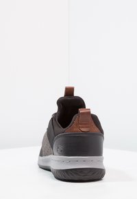 Sneaker nera con texture perforata, dettagli in pelle marrone e suola in gomma grigia. Presenta una linguetta per indossarla facilmente.