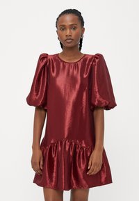 Κοντό κόκκινο σατέν φόρεμα με φουσκωτά μανίκια, στρογγυλή λαιμόκοψη και ρυάκι στο τελείωμα. Το ύφασμα έχει γυαλιστερή επιφάνεια και απαλό υφή.