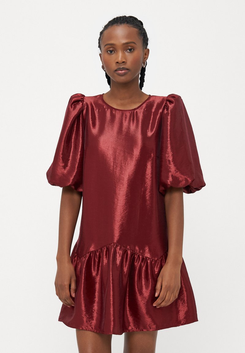 Κοντό κόκκινο σατέν φόρεμα με φουσκωτά μανίκια, στρογγυλή λαιμόκοψη και ρυάκι στο τελείωμα. Το ύφασμα έχει γυαλιστερή επιφάνεια και απαλό υφή.