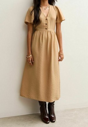Femme portant une robe midi beige avec des manches courtes volantées et des bottines marron, debout devant un mur clair uni.