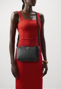 Black crossbody bag with a textured finish, silver hardware, and logo detail, worn over a red fitted dress with a square neckline.