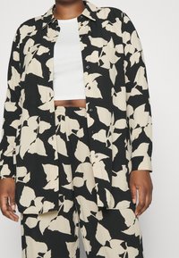 Camicia button-up nera e crema con motivo a foglie. Tessuto morbido, vestibilità comoda, bottoni visibili e maniche lunghe. Indossata sopra un top corto bianco.