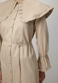 Abito beige con bottoni, dotato di un ampio colletto a volant, vita regolabile con coulisse e maniche arricciate. Tessuto liscio con una consistenza morbida.