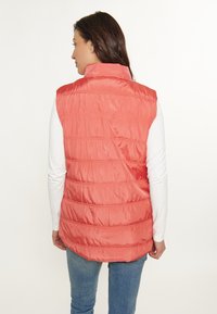 Koraalrood puffy vest met een hoge kraag en horizontale stiksels. Draag het over een witte blouse met lange mouwen. De stof lijkt glad en glanzend.