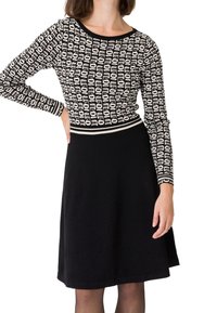 Femme portant un haut à manches longues à motifs noirs et blancs avec une jupe noire en ligne A, vue du cou aux genoux sur fond blanc.