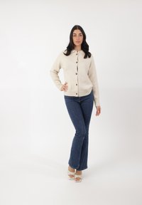 Beige gebreide cardigan met ronde kraag, voorzien van bruine knopen, gedragen over een zwarte top en gecombineerd met blauwe flared jeans en witte sandalen.