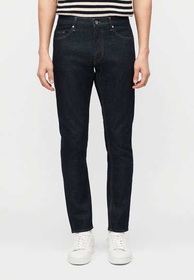 Persona che indossa jeans aderenti scuri, top a righe bianco e nero e sneakers bianche, in piedi contro uno sfondo chiaro e uniforme.