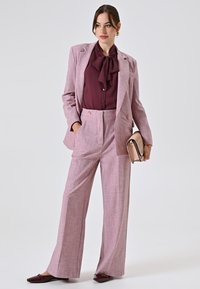 Giacca e pantaloni su misura rosa chiaro con un motivo a quadri, abbinati a una blusa bordeaux scuro e a una borsa rosa chiaro.