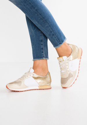 Füße in blauen Jeans und weißen Sneakers mit goldenen Akzenten, die auf einem Fuß stehen, während der andere leicht angehoben ist.