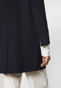 Navy coat made of a textured material with a straight cut, featuring a tailored sleeve. A cream ribbed sweater peeks from underneath.