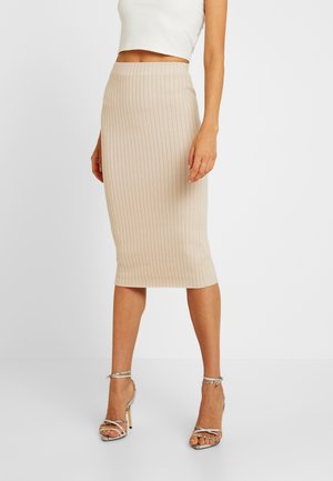 Kvinde iført en beige ribstrikket midi nederdel kombineret med en hvid cropped ærmeløs top og sølvfarvede stroppesandaler med høje hæle.