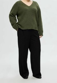 Maglione verde oliva con scollo a V in tessuto texture e orlo grezzo abbinato a pantaloni larghi neri, caratterizzati da una trama liscia e un design semplice.