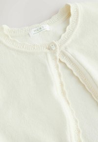 Cardigan en tricot couleur crème avec bords à volants, fermeture à un bouton blanc, et étiquette "NEXT Made in Bangladesh" à l'intérieur du col.