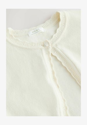 Krémový pletený sveter na gombíky s volánikovými okrajmi, zapínanie na jeden biely gombík a štítok "NEXT Made in Bangladesh" vo vnútri goliera.