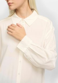 Weißes, leichtes, langärmliges Hemd mit Button-Down-Kragen; verfügt über eine glatte Textur und gemuffte Ärmel mit Knopfakzenten.