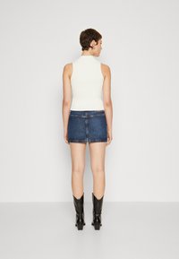 Hvit ermløs topp kombinert med et denim miniskjørt. Modellen har på seg svarte knelange støvler. Glatt tekstur med en tettsittende silhuett.