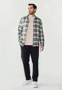 Καρό πουκάμισο με κουμπιά σε πράσινο, ναυτικό μπλε και λευκό, φορεμένο πάνω από μπλούζα μπεζ με στρογγυλή λαιμόκοψη. Στενά μαύρα παντελόνια και λευκά αθλητικά παπούτσια.