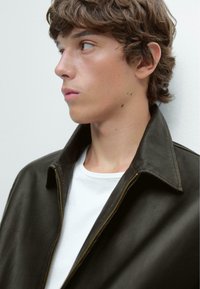Brown leather jacket with a zipper and wide collar, worn over a white T-shirt. Smooth texture, fitted design, and minimalistic style.