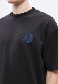 Μαύρο βαμβακερό T-shirt με στρογγυλή λαιμόκοψη, που διαθέτει μπλε κεντητό κυκλικό λογότυπο στην αριστερή πλευρά του στήθους. Κοντά μανίκια.