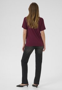 T-shirt bourgogne à manches courtes associé à un jean noir droit. Le modèle porte des mules à talons noirs, avec des cheveux longs et ondulés visibles.