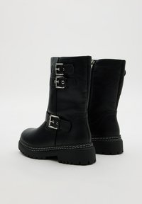 Botas de piel negras hasta el tobillo con dos hebillas de plata, una superficie texturizada y una suela de goma negra gruesa con un patrón de relieve.