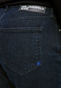 Os jeans de denim azul escuro apresentam uma etiqueta preta proeminente com texto branco na cintura e um logótipo bordado azul no bolso traseiro.
